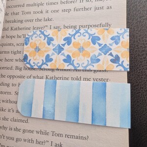 Peut inclure: Deux marque-pages décoratifs reposent sur une page de livre. L'un présente un motif floral bleu et jaune, l'autre des rayures verticales bleues et blanches. Les deux sont rectangulaires avec des coins arrondis, parfaits pour marquer les pages.