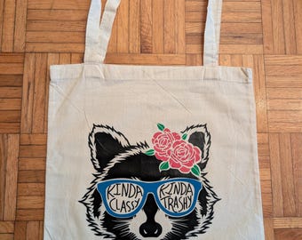 May include: A white canvas tote bag featuring a black and white raccoon wearing blue sunglasses and a pink flower crown. The sunglasses read "Kinda Classy" and "Kinda Trashy".