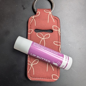 May include: A red lip balm holder with a silver key ring and a grape-flavored lip balm. The holder has a white bow pattern. The lip balm is purple and white, with the text "LIP BUTTER" and "GRAPE".
