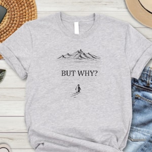 May include: A light gray t-shirt with the text "BUT WHY?" above a small penguin illustration. The shirt is paired with blue jeans, black and white sneakers, sunglasses, a smartphone, a straw woven placemat, a straw hat, and a potted plant.