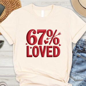 May include: Cream-colored t-shirt with the text "67% LOVED" in red, featuring hearts and an arrow. The shirt is styled with blue jeans, a straw mat, sunglasses, a phone, a hat, and sneakers.
