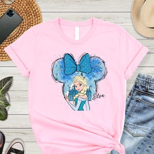 May include: Pink t-shirt with a blue and white illustration of Elsa from Frozen wearing a blue bow and standing in front of a castle. The illustration is in the shape of Mickey Mouse ears. The text "Elsa" is written below the illustration.