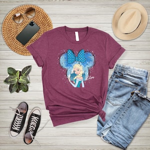 May include: A maroon t-shirt with a blue and white graphic of Elsa from Frozen. The graphic features Elsa with a blue bow and a castle in the background. The text "Elsa" is printed below the graphic.