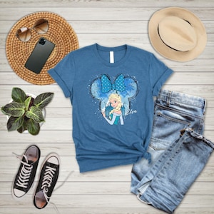 May include: A blue t-shirt with a graphic of Elsa from Frozen in a Mickey Mouse ear silhouette with a blue bow. The text "Elsa" is printed below the graphic.
