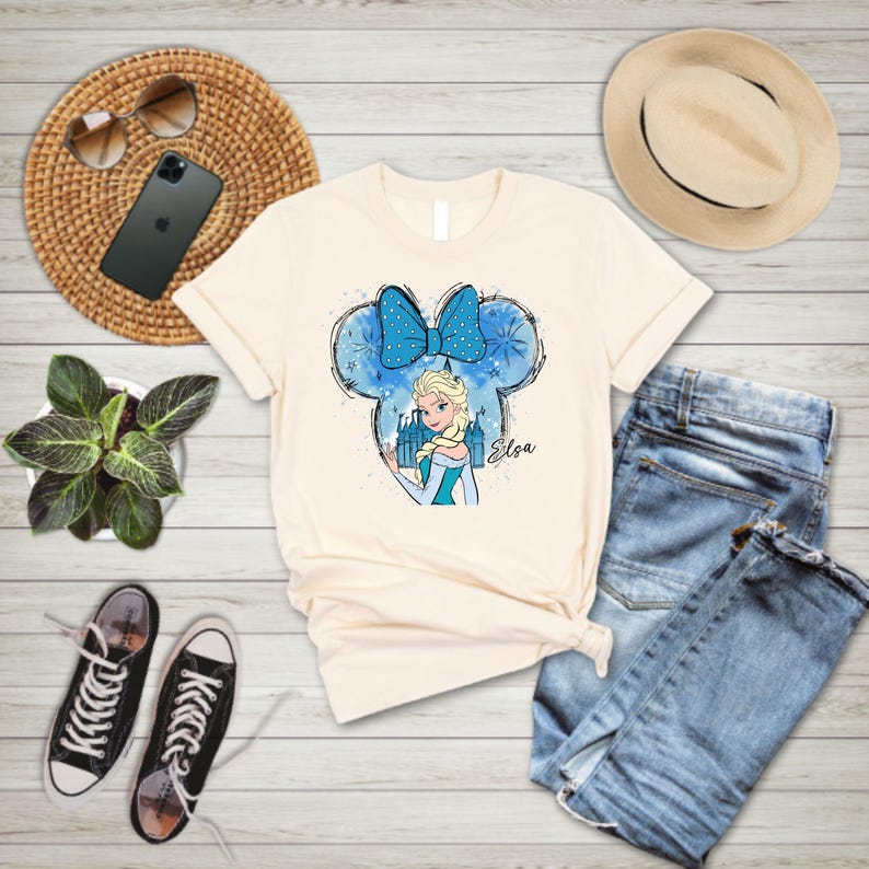 May include: A light beige t-shirt with a blue and white graphic of Elsa from Frozen in a Mickey Mouse ear silhouette. The graphic includes a blue bow, snowflakes, and the text "Elsa".