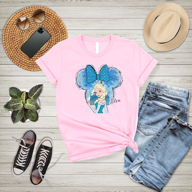 May include: Pink t-shirt with a blue and white illustration of Elsa from Frozen wearing a blue bow and standing in front of a castle. The illustration is in the shape of Mickey Mouse ears. The text "Elsa" is written below the illustration.