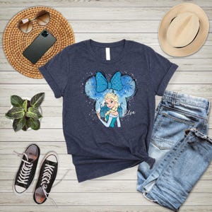 May include: A navy blue t-shirt with a graphic of Elsa from Frozen in a blue and white dress with a blue bow. The graphic is in a Mickey Mouse ear shape with snowflakes and the word "Elsa" written below.