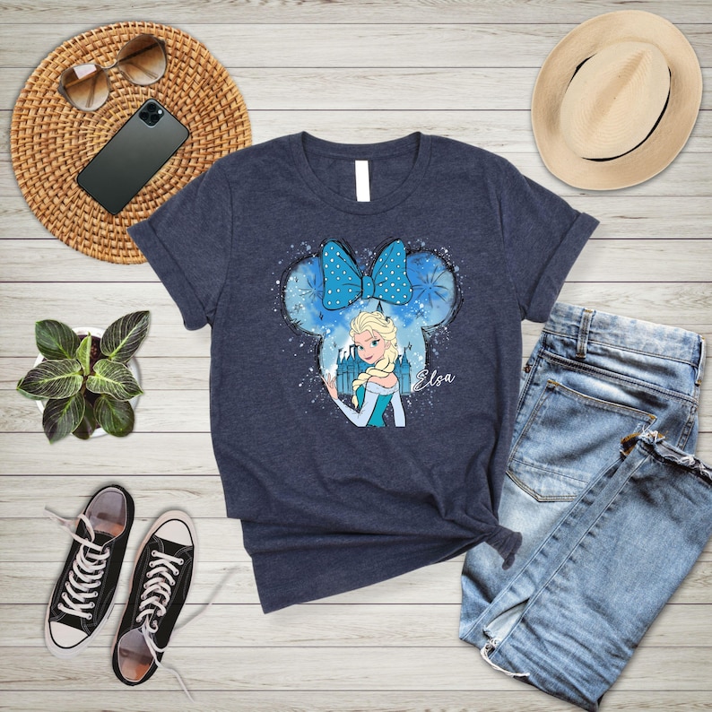 May include: A navy blue t-shirt with a graphic of Elsa from Frozen in a blue and white dress with a blue bow. The graphic is in a Mickey Mouse ear shape with snowflakes and the word "Elsa" written below.