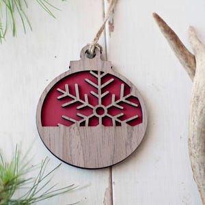 May include: A wooden Christmas ornament in the shape of a ball with a red snowflake cutout. The ornament is made of wood with a natural finish and has a rustic look.