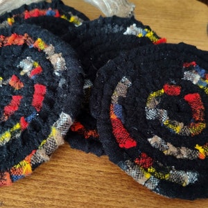 May include: A set of four black, circular coasters with a spiral design. The spiral is made of colorful fabric scraps in red, yellow, blue, and brown. The coasters are handmade and have a rustic, textured appearance.