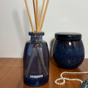 Könnte beinhalten: Ein blauer Glas-Diffusor für ätherische Öle mit Holzstäbchen, ein kleiner blauer Keramiktopf und eine Perlenkette mit einem türkisfarbenen Vogelanhänger. Auf dem Diffusor steht das Wort "sensen".