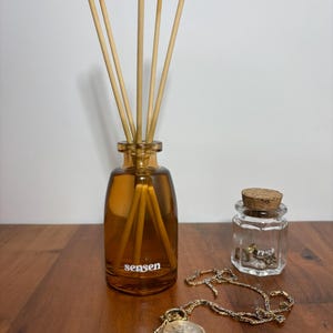 Może przedstawiać: Bursztynowa szklana butelka z dyfuzorem i drewnianymi patyczkami, z napisem "sensen", stoi na drewnianej powierzchni. Obecny jest również mały szklany słoik z korkiem i złoty zegarek kieszonkowy z łańcuszkiem. Scena sugeruje zapach do domu i akcesoria w stylu vintage.