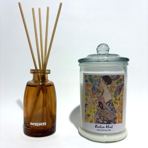 Può includere: Un diffusore in vetro ambrato con bastoncini di legno e la scritta "sensen" stampata sul davanti. Accanto, una candela in barattolo di vetro con coperchio, con una stampa di "Lady with Fan, 1918" di Gustav Klimt.