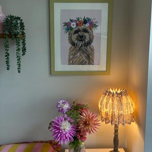 May include: A framed print of a dog wearing a floral crown hangs on a light gray wall. A table lamp with a patterned shade sits on a white table. A vase of flowers and a striped pillow are also visible.