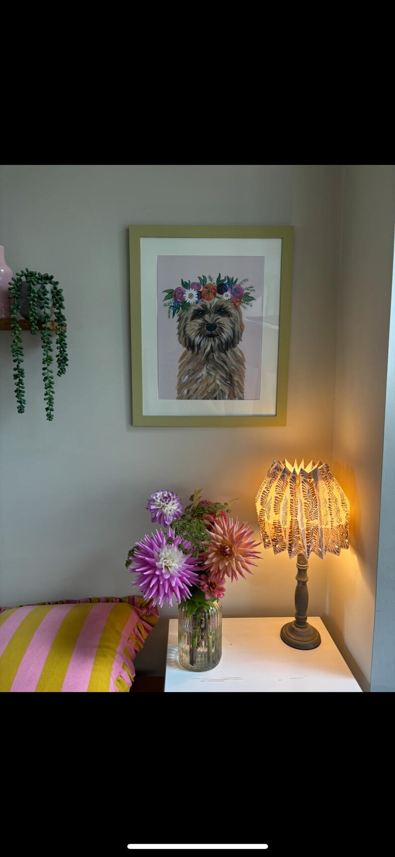 May include: A framed print of a dog wearing a floral crown hangs on a light gray wall. A table lamp with a patterned shade sits on a white table. A vase of flowers and a striped pillow are also visible.