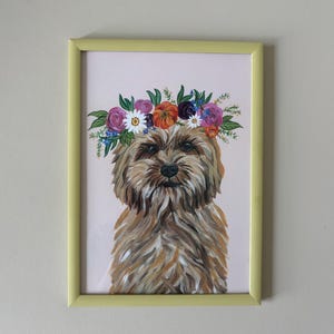 May include: A framed painting of a dog adorned with a floral crown. The dog has brown and tan fur and a dark nose. The floral crown features purple, orange, and white flowers. The frame is a pale yellow, and the background is a pale pink.