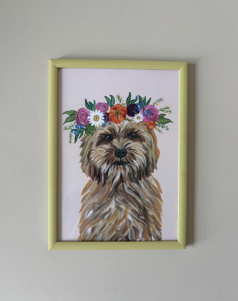 May include: A framed painting of a dog wearing a floral crown. The dog has brown and tan fur and a dark nose. The floral crown features purple, orange, and white flowers. The frame is a light yellow color, and the background is a pale pink.