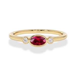 Op de afbeelding: Een gouden ring met een ovale robijn in het midden en twee kleinere, ronde, heldere edelstenen. De ring heeft een eenvoudig, elegant ontwerp. De edelstenen zijn gezet in een gepolijste gouden band. Het ontwerp van de ring is klassiek.