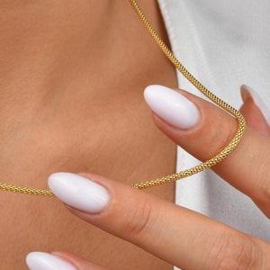 May include: A delicate gold necklace with a textured chain is held by a person. The necklace is a bright gold colour and appears to be made of small, interconnected links. The person is wearing a white top.