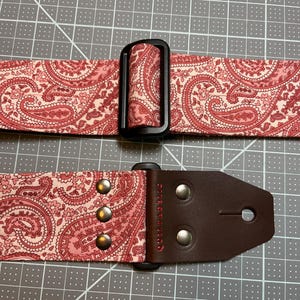 May include: A guitar strap with a pink paisley pattern. The strap has a black plastic buckle and a brown leather end piece with silver rivets. The word "Quiltaholic" is printed on the leather.