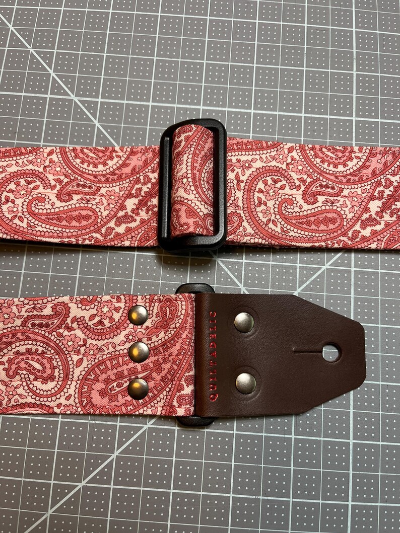 May include: A guitar strap with a pink paisley pattern. The strap has a black plastic buckle and a brown leather end piece with silver rivets. The word "Quiltaholic" is printed on the leather.