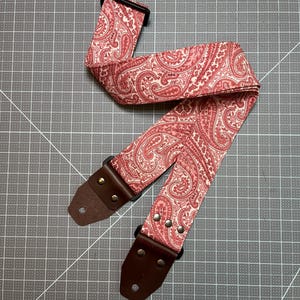 May include: A guitar strap with a paisley pattern in shades of red and cream. The strap has brown leather ends with metal rivets and a black plastic adjustment buckle. The strap is designed for use with an electric or acoustic guitar.