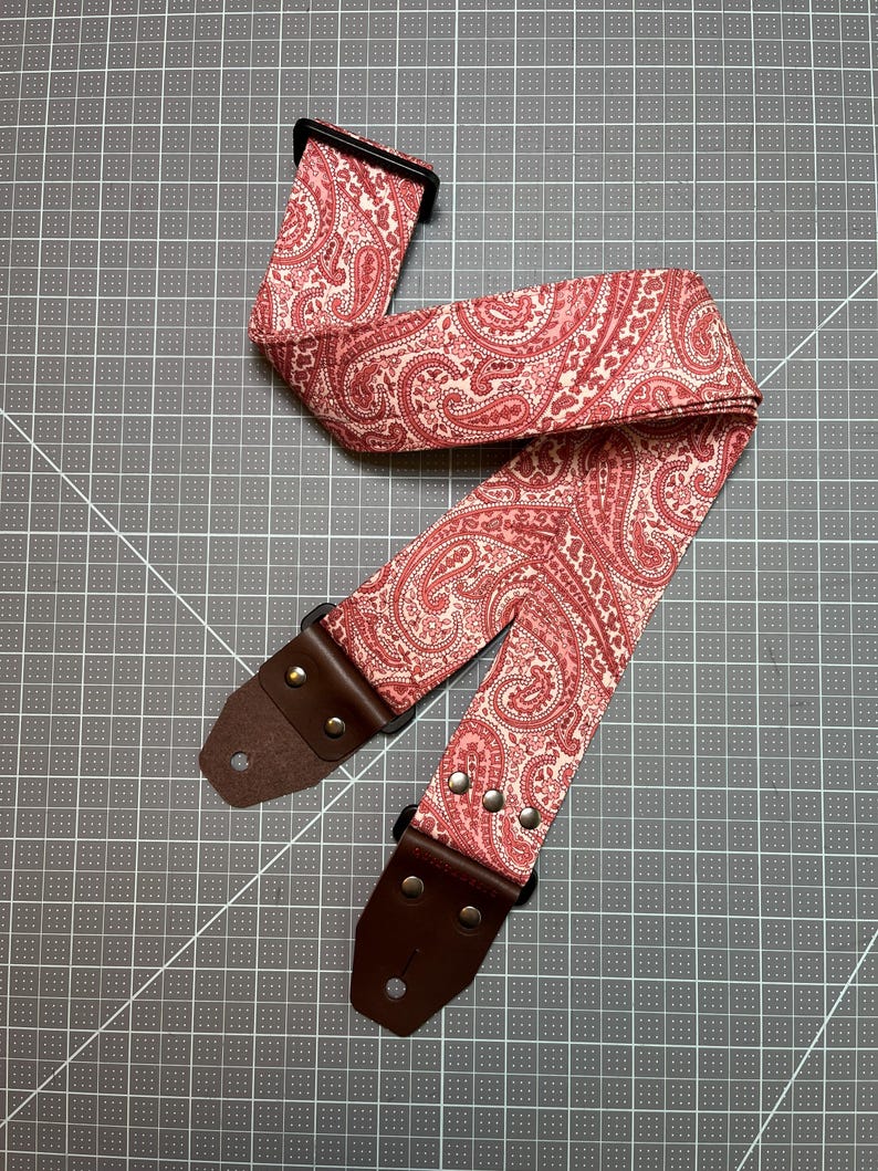 May include: A guitar strap with a paisley pattern in shades of red and cream. The strap has brown leather ends with metal rivets and a black plastic adjustment buckle. The strap is designed for use with an electric or acoustic guitar.