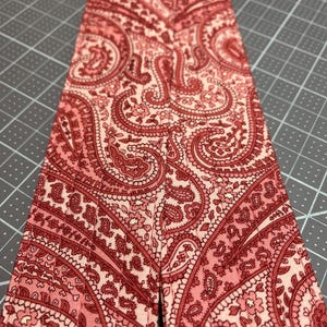 May include: Close-up of a fabric strip with a paisley pattern in shades of red and pink. The intricate design features floral and curved elements. The fabric is likely cotton or a similar material, suitable for sewing or crafting projects.