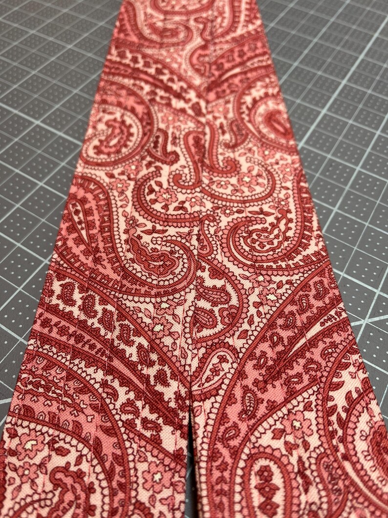 May include: Close-up of a fabric strip with a paisley pattern in shades of red and pink. The intricate design features floral and curved elements. The fabric is likely cotton or a similar material, suitable for sewing or crafting projects.