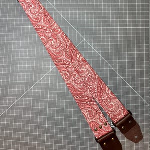 May include: A guitar strap with a red and white paisley pattern. The strap has brown leather ends and metal hardware. The strap is designed to be worn over the shoulder and is adjustable.