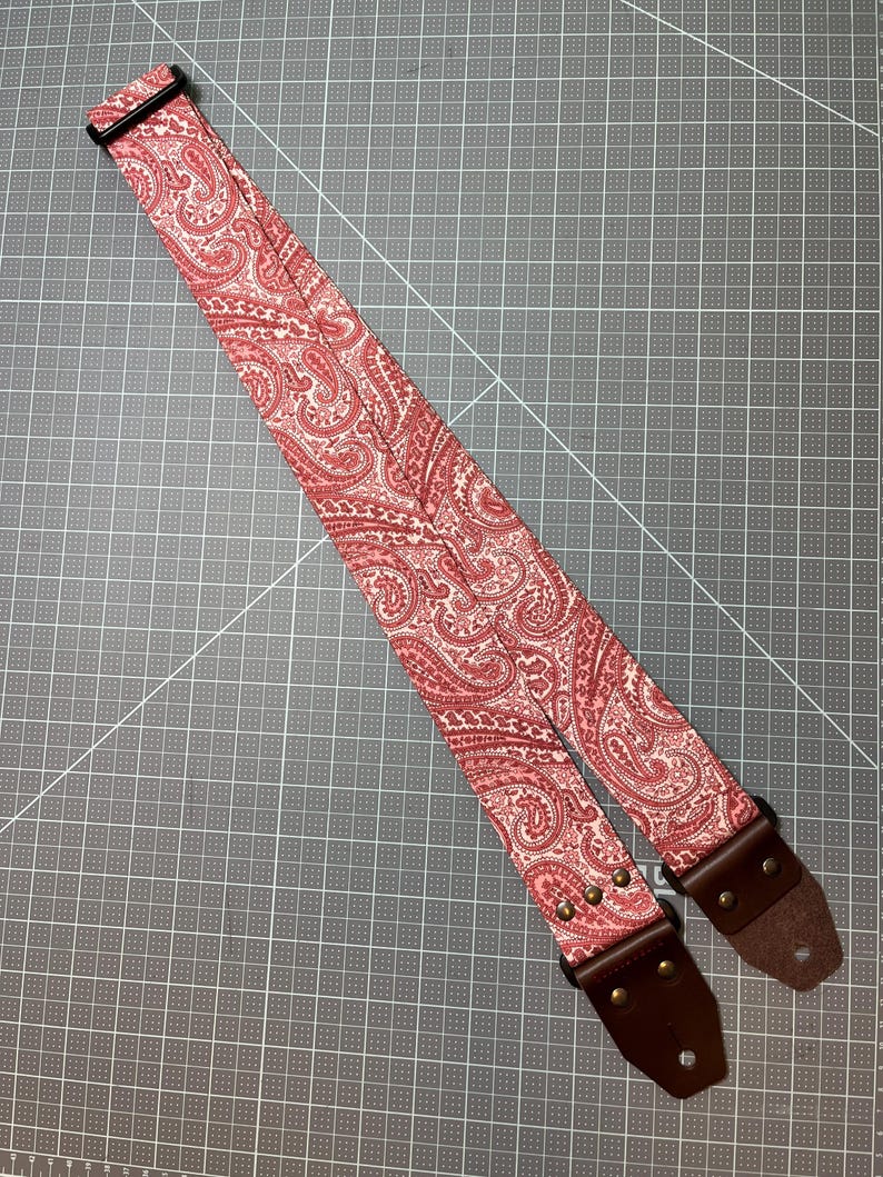 May include: A guitar strap with a red and white paisley pattern. The strap has brown leather ends and metal hardware. The strap is designed to be worn over the shoulder and is adjustable.