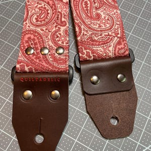 May include: A guitar strap with a paisley pattern in shades of pink and red. The strap has brown leather ends secured with silver rivets. The text "QUILTADELIC" is embroidered in red on one end. The strap is designed for musical instruments.