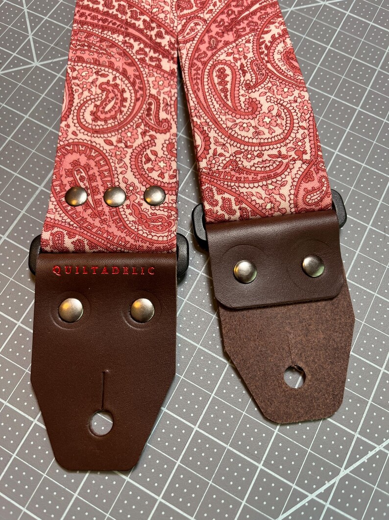 May include: A guitar strap with a paisley pattern in shades of pink and red. The strap has brown leather ends secured with silver rivets. The text "QUILTADELIC" is embroidered in red on one end. The strap is designed for musical instruments.