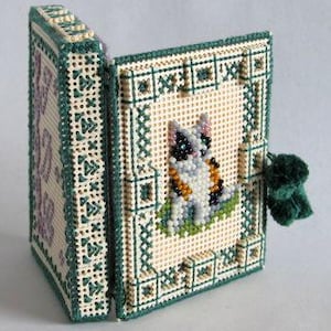 Puede incluir: Un pequeño libro de punto de aguja verde y blanco con un diseño de gato. El gato es blanco con marcas negras y grises y está sentado en un parche de hierba verde. El libro tiene un borla verde.