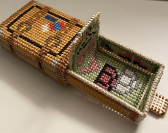 May include: A handmade, open needlepoint box with a hinged lid. The box is primarily tan with black, white, red, and blue accents. The interior features a green, pink, and white design. The box is likely used for storing small items.