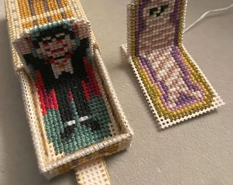 May include: Two small, hand-stitched coffins with a gold trim. One coffin is open and contains a cross-stitch of a vampire. The other coffin is closed and has a cross-stitch of a mummy inside.