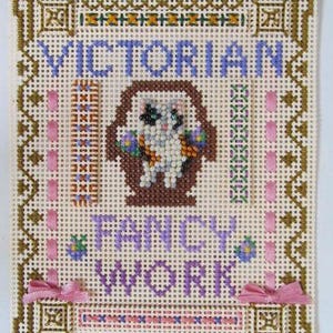 Könnte beinhalten: Stickerei im viktorianischen Stil mit einer Katze in einem Korb. Der Text ist in Lila und Blau gehalten, mit einem braunen Rand und rosa Schleifen. Die Katze ist weiß, schwarz und orange.