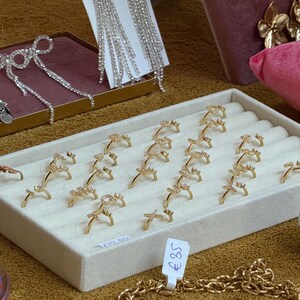 May include: A white display tray with 18 gold rings with clear stones. The rings are arranged in rows and are all the same design. The tray is on a brown surface. The price tag reads "&pound;35".