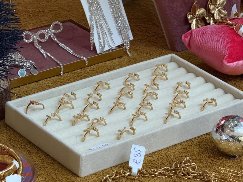 May include: A white display tray with 18 gold rings with clear stones. The rings are arranged in rows and are all the same design. The tray is on a brown surface. The price tag reads "&pound;35".