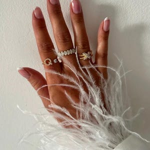 May include: A hand with white french manicured nails wearing several gold rings with small diamonds. The hand is resting on a white fluffy feather boa.