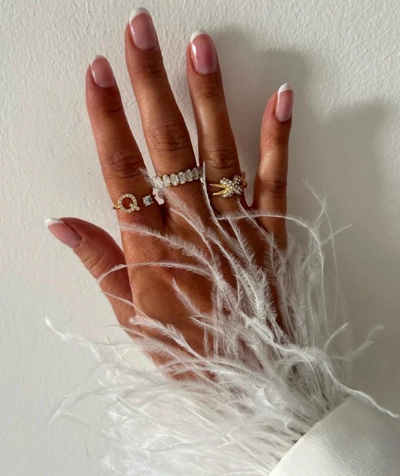 May include: A hand with white french manicured nails wearing several gold rings with small diamonds. The hand is resting on a white fluffy feather boa.