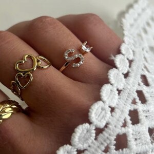May include: A gold ring with a single letter 'S' set with small clear stones. The ring is on a finger wearing a gold ring with a chain of hearts.