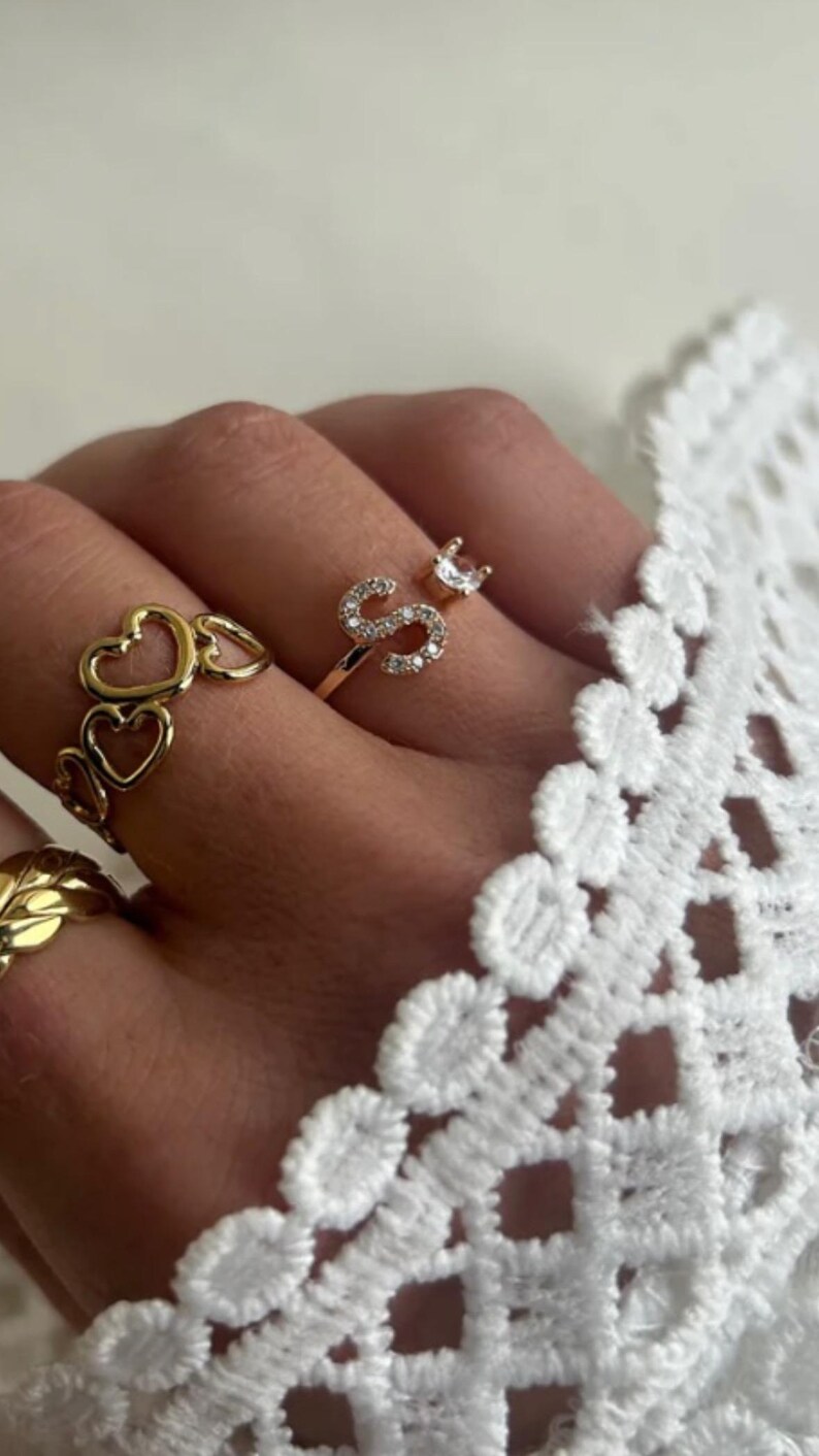 May include: A gold ring with a single letter 'S' set with small clear stones. The ring is on a finger wearing a gold ring with a chain of hearts.