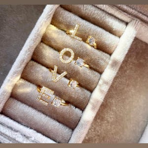 May include: A set of four gold rings with clear stones spelling out the word "LOVE". The rings are displayed in a velvet ring box.