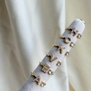 May include: A set of gold rings with clear cubic zirconia stones, each ring features a different letter of the alphabet. The rings are displayed on a white velvet stand.