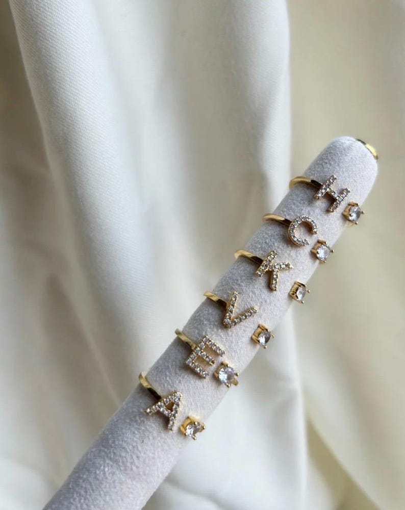May include: A set of gold rings with clear cubic zirconia stones, each ring features a different letter of the alphabet. The rings are displayed on a white velvet stand.