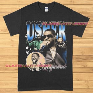 Può includere: Una maglietta nera con una grafica in stile vintage di Usher Raymond. La grafica presenta un ritratto di Usher con il testo "Usher Raymond" in un gradiente blu e bianco.