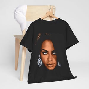 May include: Black t-shirt with a portrait of a woman with dark hair and large, sparkling earrings. The image is a close-up of the woman's face, with a focus on her eyes and lips. A white baseball cap rests on a wooden chair.