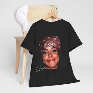 May include: Black t-shirt with a portrait of a smiling person wearing a brown bandana. The image includes a white signature. A white baseball cap rests on a wooden chair in the background.