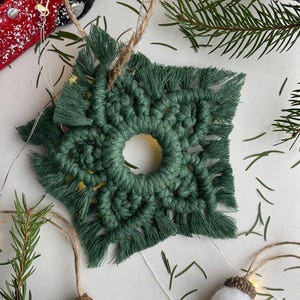 May include: A handmade green macrame snowflake ornament with a central hole, surrounded by fringed edges. Accompanying the ornament are small acorn-shaped decorations with white pom-poms and brown caps, all on a white surface with pine needles.